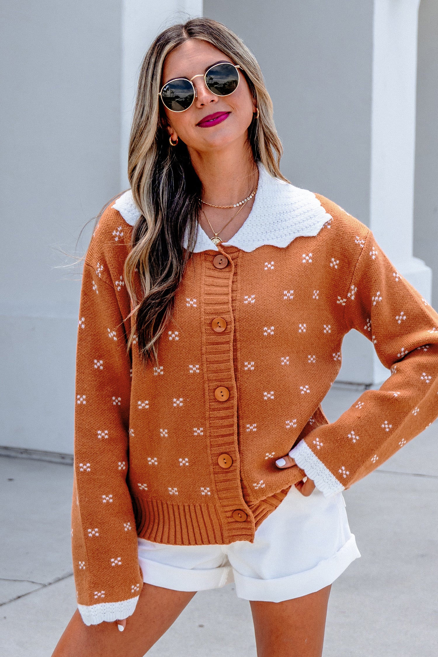 A woman wearing a rust-orange knit cardigan with a white scalloped collar and white button details, paired with white shorts and sunglasses, posing outdoors against a light gray wall.