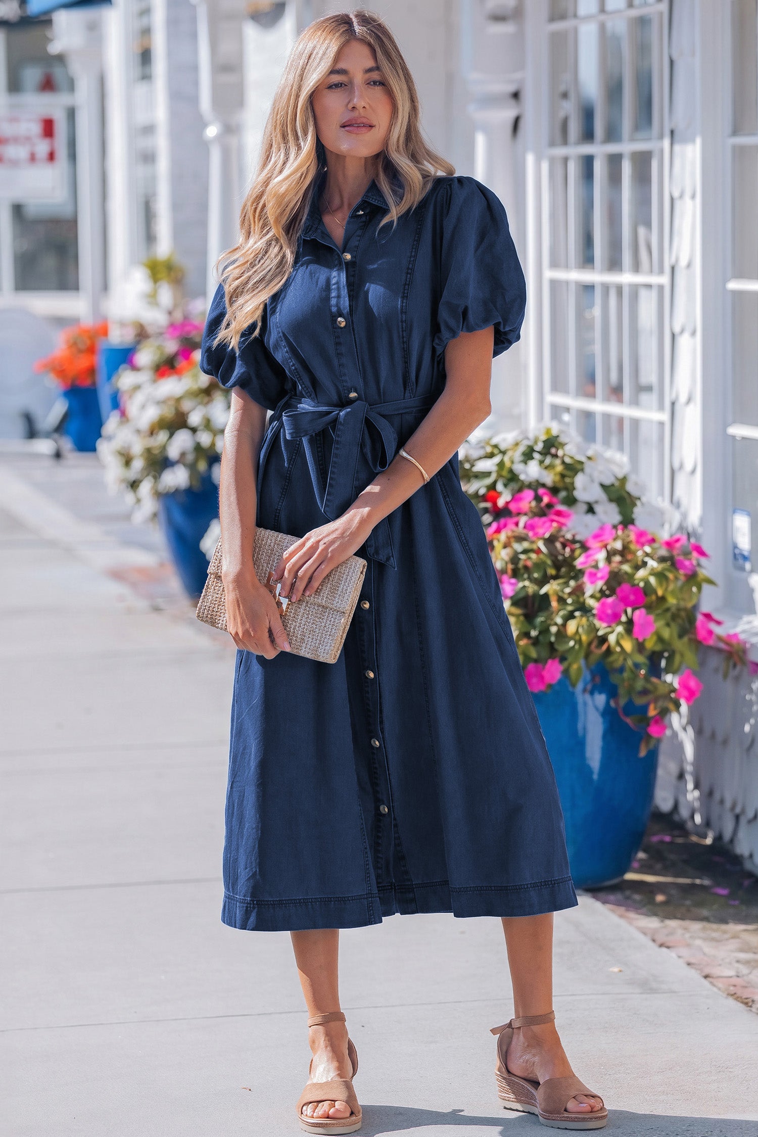 Woman wearing a blue dress standing on a sidewalk with flowers and buildings in the background