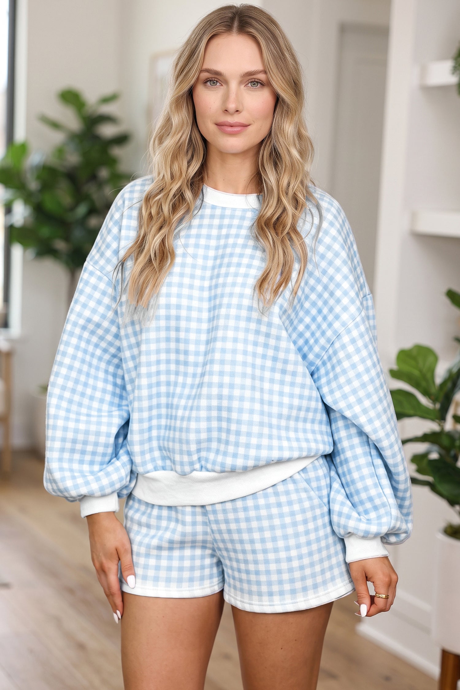 A woman modeling a light blue and white gingham lounge set, featuring a long-sleeve sweatshirt with balloon sleeves and matching shorts, in a bright indoor setting.