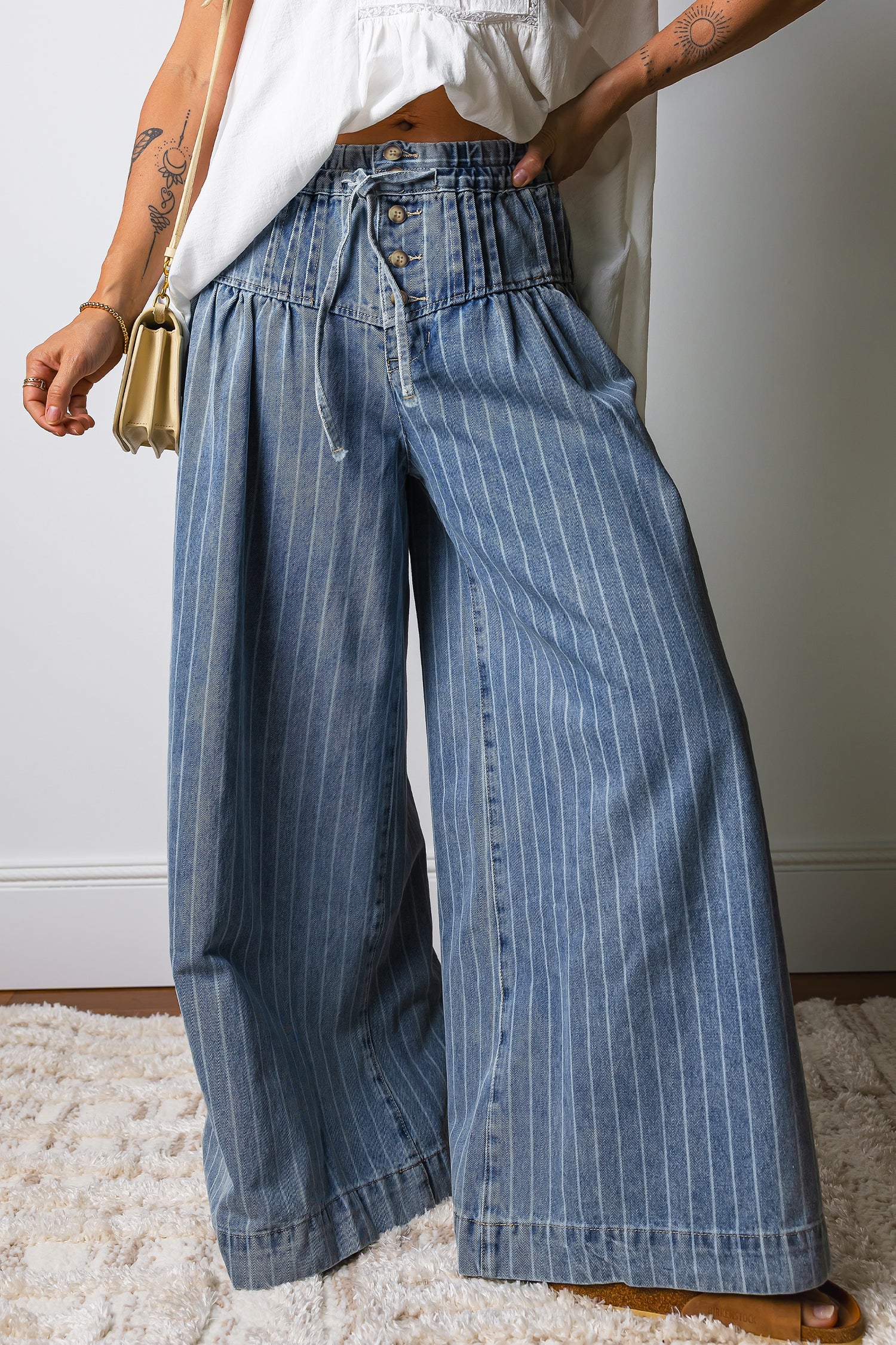 A person wearing wide-leg blue denim jeans with thin white vertical pinstripes, styled with a white top and a small gold handbag, standing on a white shag rug.