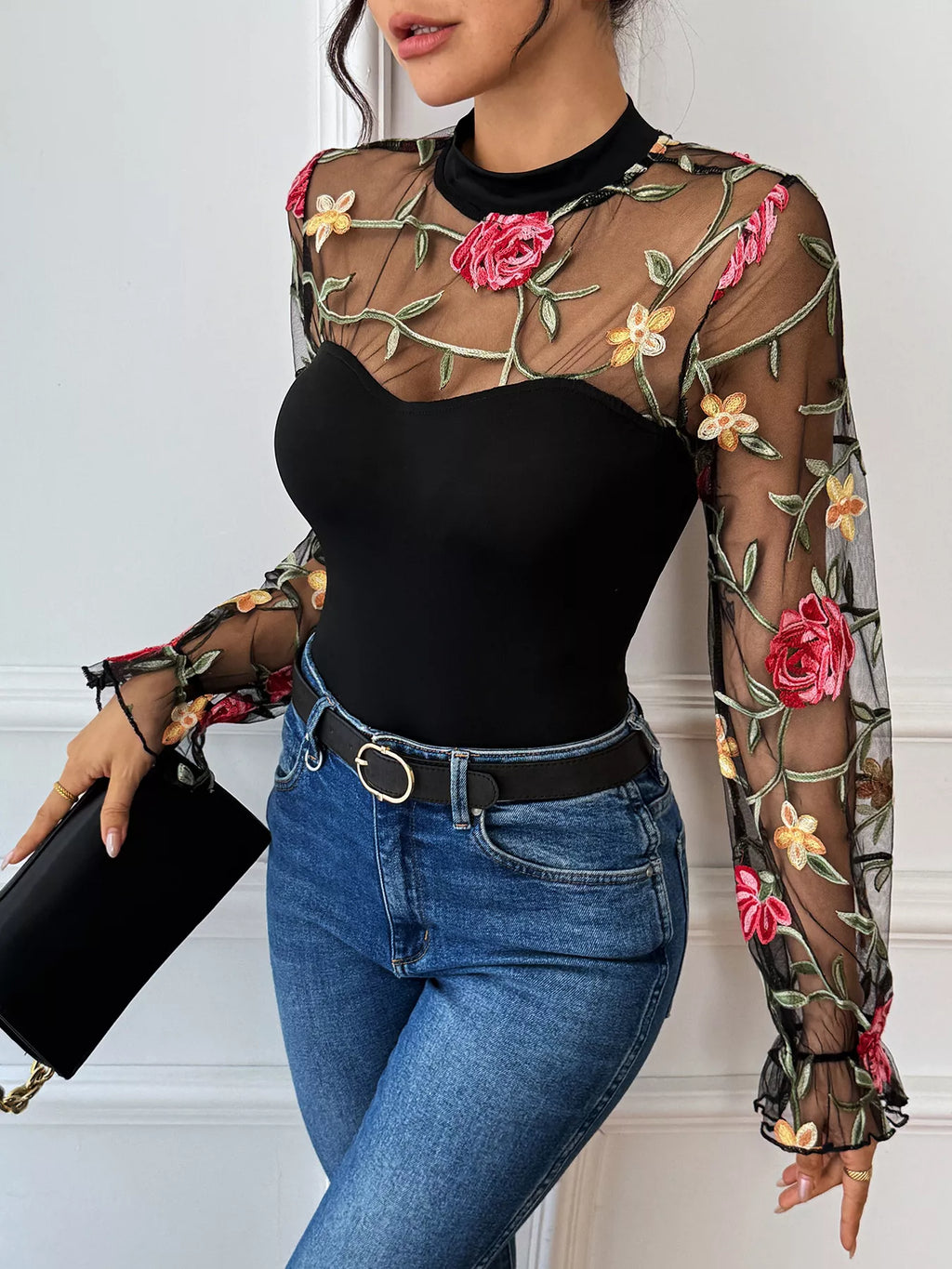 Person wearing a black bodysuit with floral embroidery and blue jeans, holding a black clutch-DEA Collection 