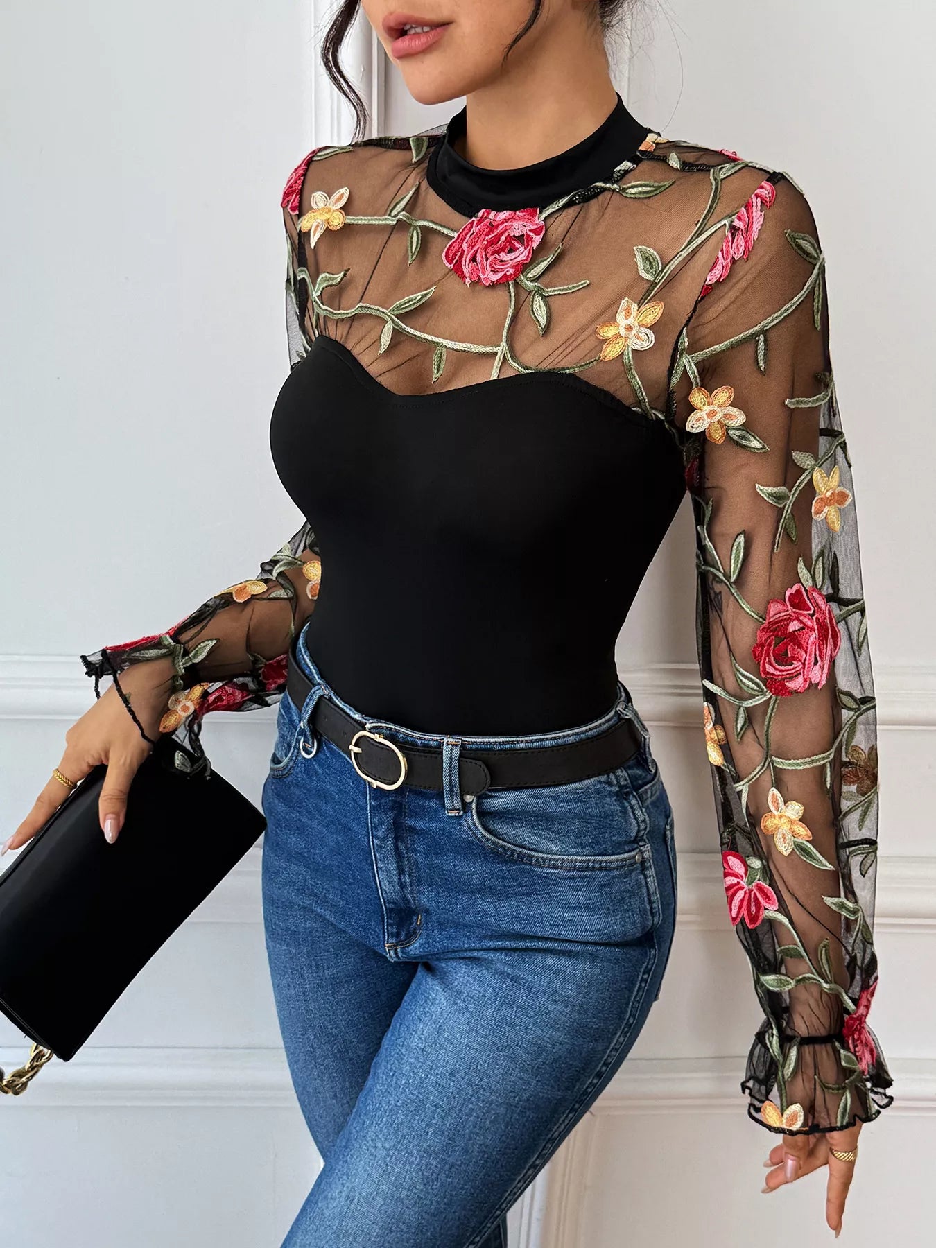 Person wearing a black bodysuit with floral embroidery and blue jeans, holding a black clutch-DEA Collection 