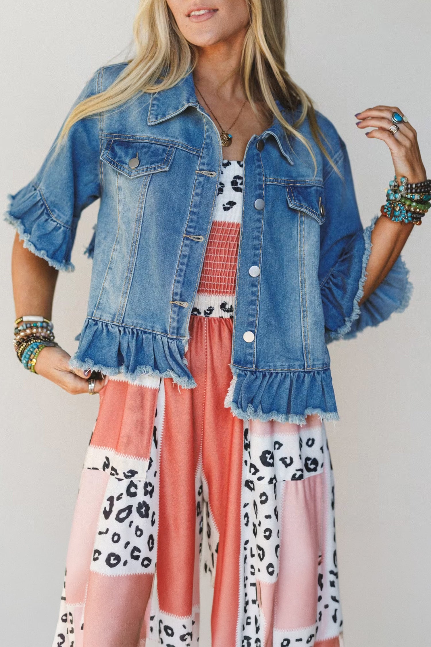 A woman wearing a light blue denim jacket with ruffled sleeves and hem, layered over a white top with pink and red patterned fabric, paired with pink patchwork wide-leg pants with black animal prints.