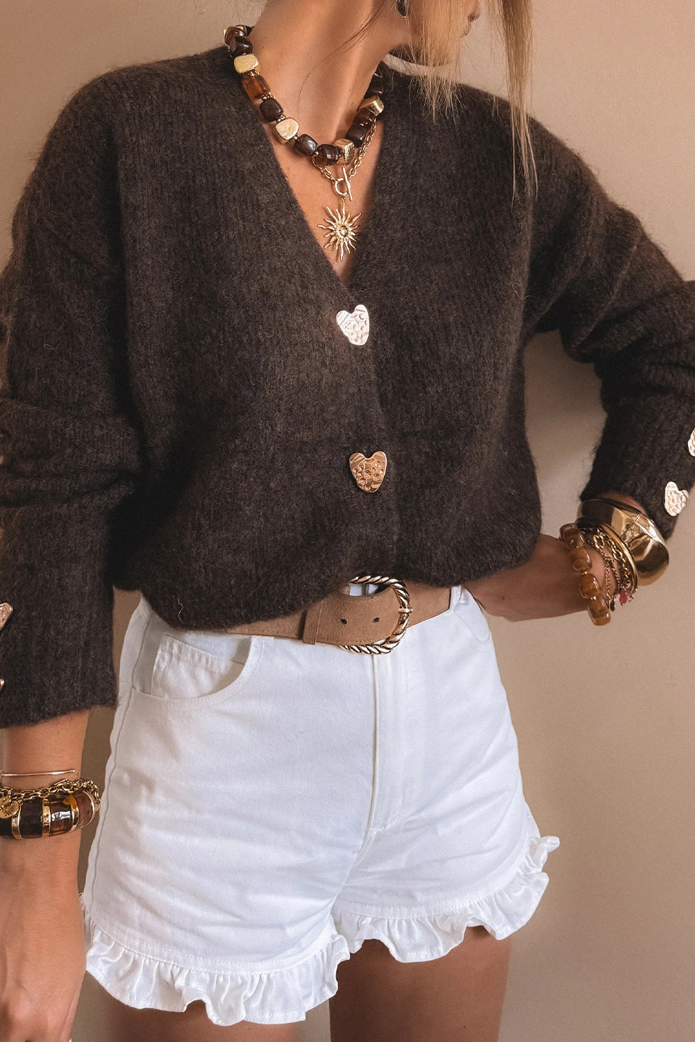 Person wearing a brown cardigan with heart buttons, white shorts, and multiple necklaces on a beige background
